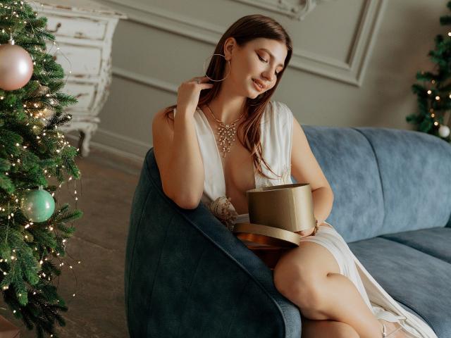 AmandaGasertoe sits on a blue velvet sofa, holding a gold gift box on a wooden tray. She has long brown hair, wears a white dress with a deep neckline, and a statement necklace. She touches her hair with her right hand, smiling softly. A decorated Christmas tree with pink and green ornaments and fairy lights is to her left. The room has a white, ornate wall panel and wooden floor. The composition is warm and festive.