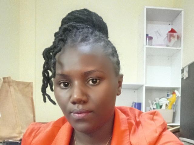 BetsiFrankie with dark skin, medium-sized tits, black dreadlocks in a high bun, wearing an orange shirt, serious expression, in a room with a white shelf.