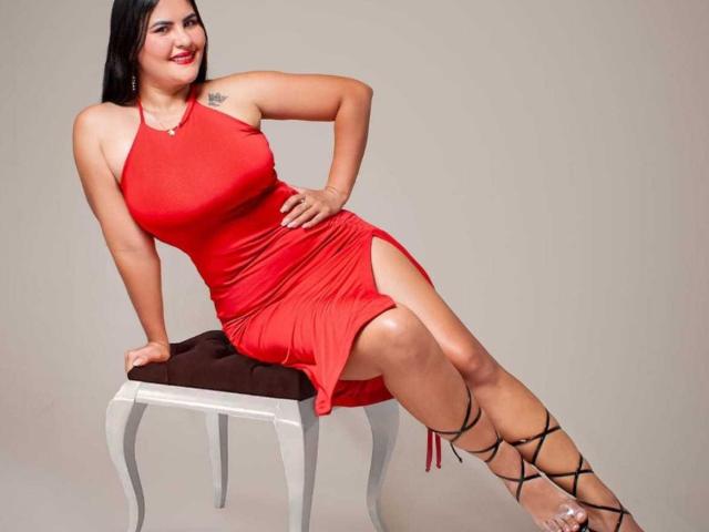 Sharlok with black hair, red dress, tits visible, sitting on white chair with brown cushion, legs crossed, black lace-up heels, tattoo on left shoulder, smiling.