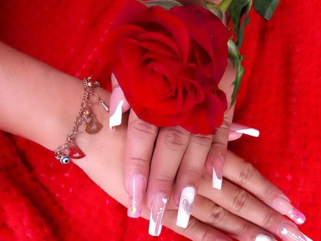 Sharlok's hands with long white and pink nails, holding a red rose, wearing a pink bracelet with heart and bead detail, on a red textured cloth.