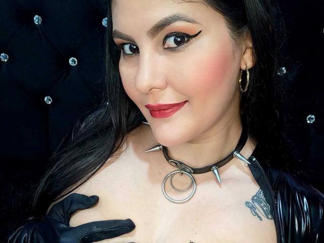 Sharlok with dark hair, red lipstick, black eyeliner, and a black choker with spikes, touching her tits with black gloves. Tattoo on right side of chest. Smirking.