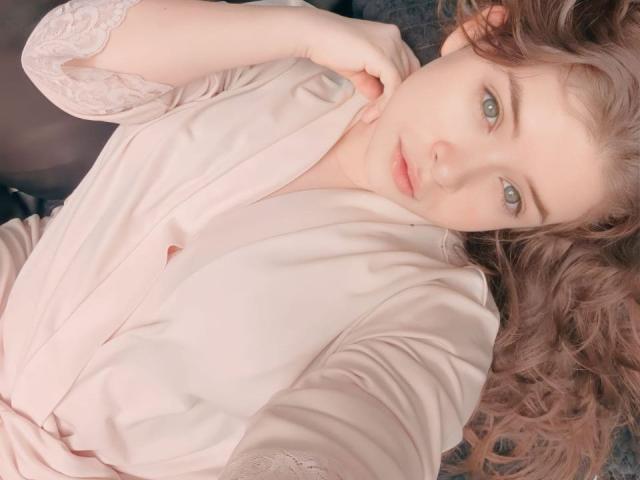 EvaRavissante with pale pink robe, light curly brown hair, and blue eyes, lying down, looking at camera, right hand touching chin, soft pink robe with lace sleeves.