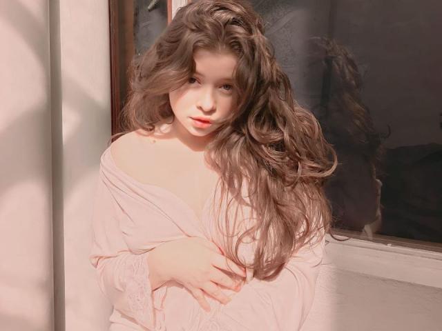 EvaRavissante with long, wavy brown hair, pale skin, and soft pink dress, standing near a window, covering her tits with one hand, looking slightly bored.