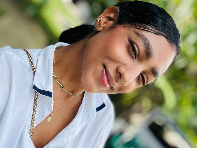 NatalyBlonder, with dark hair and tan skin, wears a white shirt, gold necklace, and earrings, smiling softly at the camera, her head tilted slightly. Her tits are partially visible.