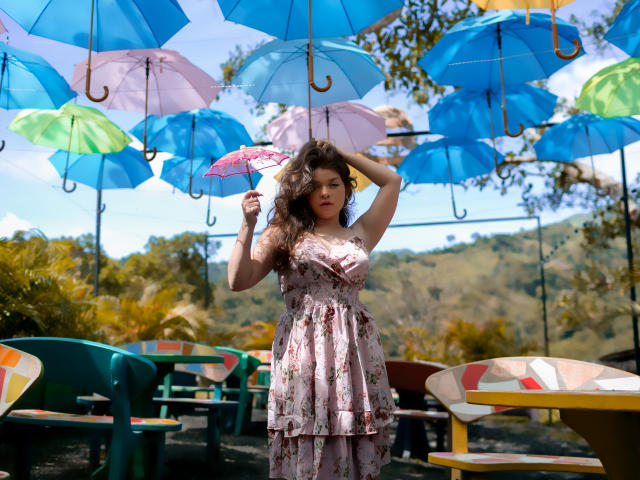 EvaRavissante with curly brown hair, wearing a floral dress, holding a pink umbrella, standing under hanging blue umbrellas, in a sunny outdoor setting with colorful tables.