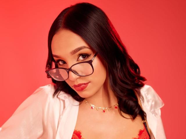 SophiaMontero with dark wavy hair, wearing glasses, red lipstick, white open shirt, red lace bra, and red necklace, looking slightly down with a subtle smile.