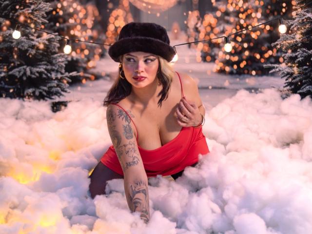 MariGonz con grandi seni, braccio tatuato, vestito rosso, cappello di pelliccia nera, inginocchiata nella neve artificiale, circondata da luci brillanti e alberi coperti di neve, che guarda la telecamera con rossetto rosso..