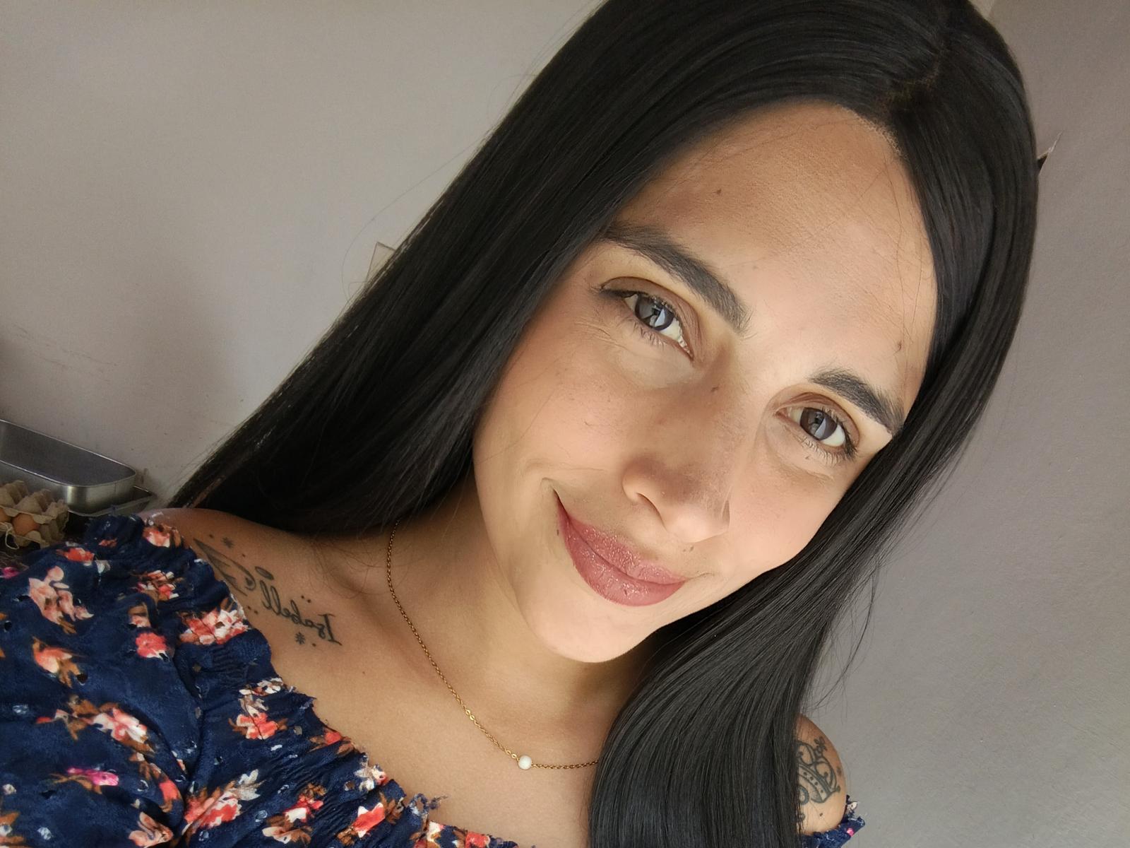 RosieFelice with long black hair, brown eyes, and a subtle smile. She's wearing a floral dress, showing a tattoo on her right shoulder. She's got small tits and a necklace.