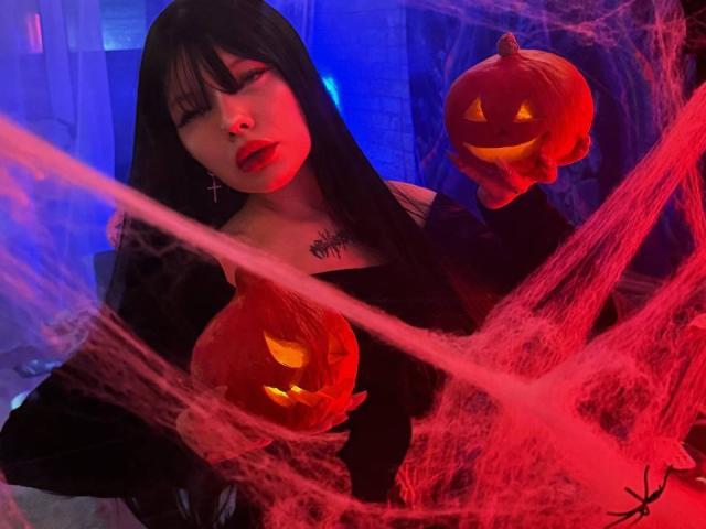 ChillOutt with black hair, red lipstick, holding two glowing jack-o'-lanterns, wearing a black off-shoulder top, red and blue light, and red spider webs in the background.