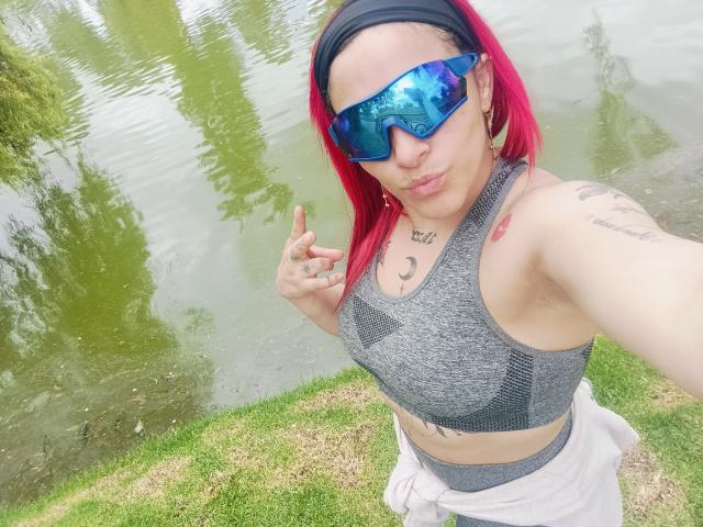 Stefanhny with red hair, blue reflective sunglasses, gray sports bra, white pants, tattoos, and a black headband, making a "rock on" gesture, near a green-grass water edge.