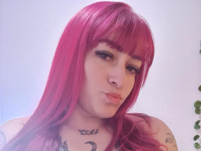 Stefanhny with pink hair, full lips, tattoos on chest and neck, and a nose ring, looking down with a slight pout. Boobs partially visible. Soft light, white background.