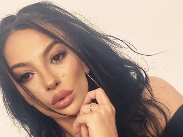 KinkyAmelie with black, wavy hair, full pink lips, dark eyebrows, and a beauty mark on her left cheek, holding her chin with a ring on her finger.