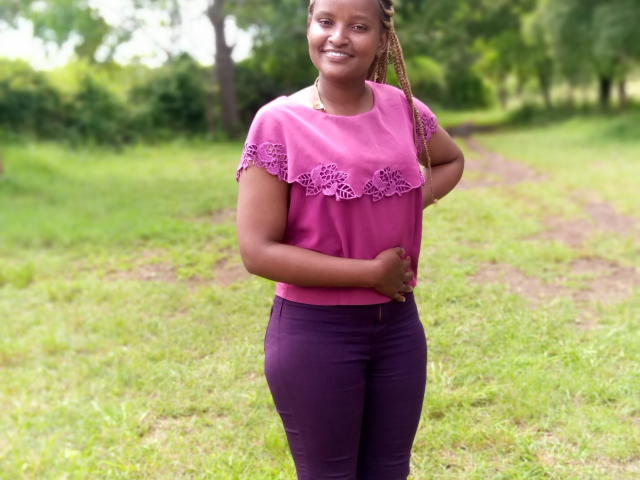 Emphi6 9 stands in a grassy area with a dirt path. She's wearing a pink lace-trimmed shirt and purple pants. Her right hand rests on her stomach, and she smiles slightly. She has braided hair. The green background includes trees and shrubbery. The colors are bright and natural, with a focus on her pink shirt and purple pants. The image has a casual, outdoor feel.