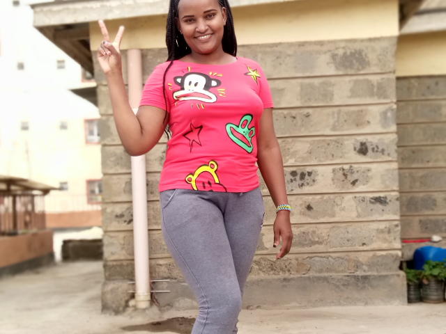 Emphi69 stands in a casual outdoor setting, smiling and flashing a peace sign with her right hand. She's wearing a bright pink t-shirt with cartoon monkey and star designs, and gray leggings. Her dark braids are visible, and she wears a green bracelet on her left wrist. The t-shirt and leggings contrast sharply, with the pink t-shirt standing out against the muted, textured stone building behind her.