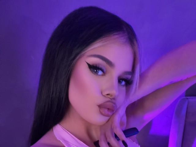 IvyRaven with long black and blonde hair, thick black eyeliner, pink blush, and pouty lips, holding a black pen near her face in purple-lit photo.