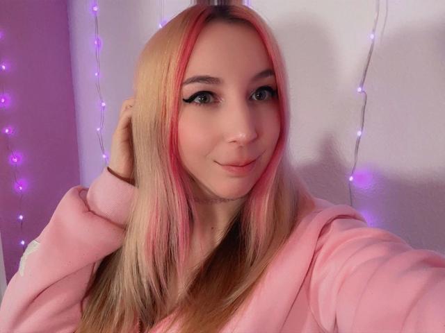 JustKerry-hot with pink and blonde hair, wearing a pink hoodie, smiling at the camera, hand in hair, with black eyeliner, in a softly lit, purple-themed photo.