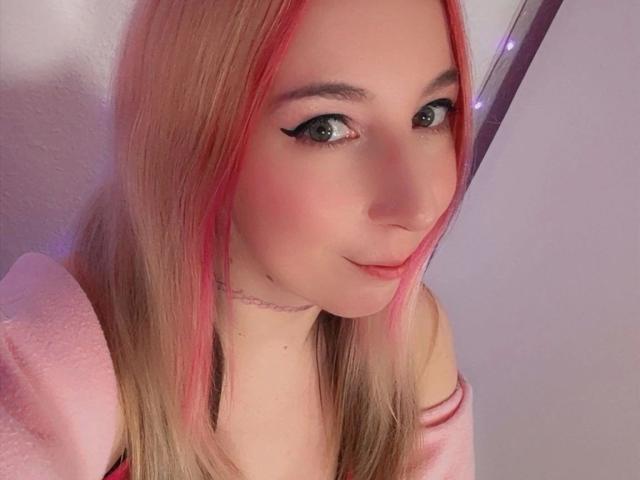 JustKerry-hot has pink-streaked blonde hair, green eyes with black eyeliner, a pink top, and a cute smile, with a slight blush on her fair skin.