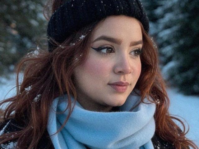 DannaXFerluccio hat rotes Haar, eine schwarze Mütze, einen blauen Schal und einen auffälligen Eyeliner. Schneeflocken befinden sich auf ihrem Gesicht und in ihrem Haar. Sie schaut zur Seite und hat ein leichtes Lächeln..