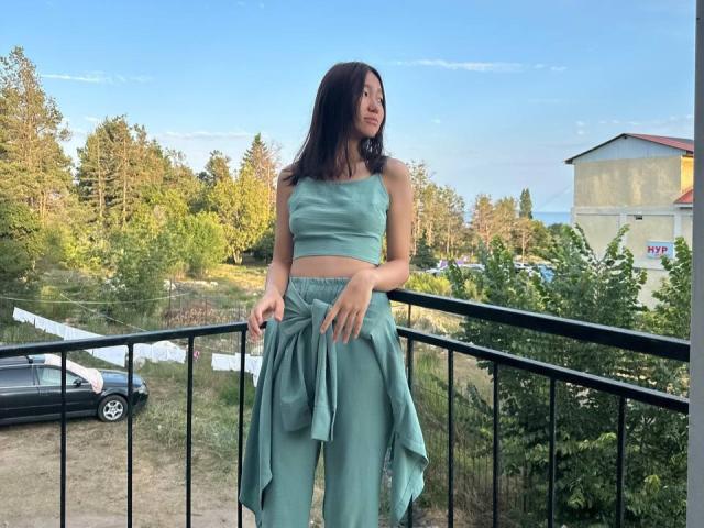 AmorBae with medium tits, wearing a green crop top and matching pants, standing on a balcony with a tied shirt, looking to the side, in a sunny outdoor setting.