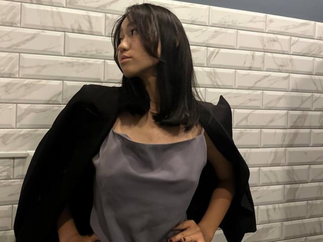 AmorBae, avec des cheveux noirs, portant une veste noire par-dessus une robe en satin gris, les mains sur les hanches, regardant sur le côté, debout devant un mur de briques blanches..
