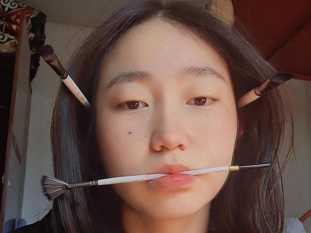 AmorBae with dark hair, two makeup brushes in hair, one in mouth, pouty pink lips, small black beauty mark on left cheek, slightly glossy skin, brown eyes.