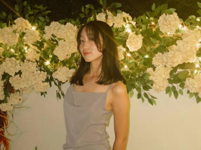 AmorBae with medium-length black hair, wearing a grey, sleeveless, cowl-neck dress, standing in front of white flowers and green leaves, illuminated by warm, yellow fairy lights.