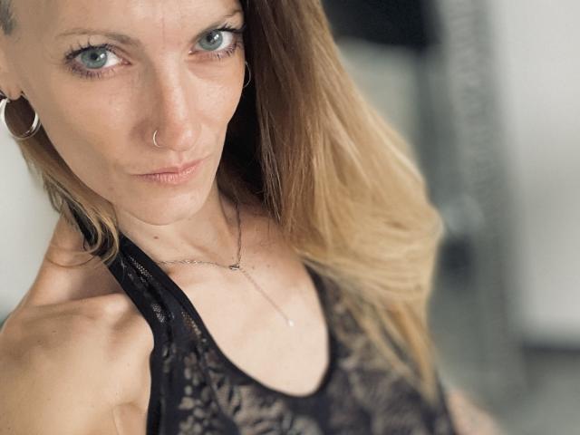 DaneeSoHot, avec ses yeux bleus, son anneau au nez et ses boucles d'oreilles, portant un haut en dentelle noire, tenant son épaule, regardant la caméra, cheveux blonds, maquillage discret, collier..