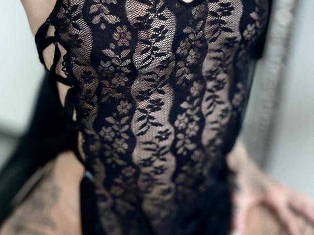 DaneeSoHot wearing a black lace bodysuit, showing her tits through the see-through fabric, with tattoos on her hand and lower arm. Her skin is light.