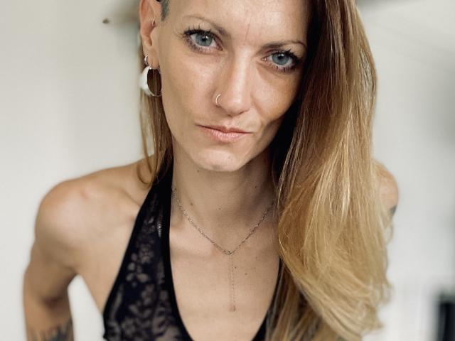 DaneeSoHot a les yeux bleus, un piercing au nez et de longs cheveux blonds. Elle porte un haut noir en dentelle avec des bretelles, un collier fin et des boucles d'oreilles cerceaux blanches, et elle a l'air sérieuse..