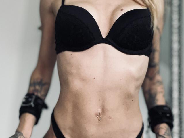 DaneeSoHot with black bra and panties, showing her tits and toned abs, tattoos on arms, black wrist cuffs, and a belly button piercing.