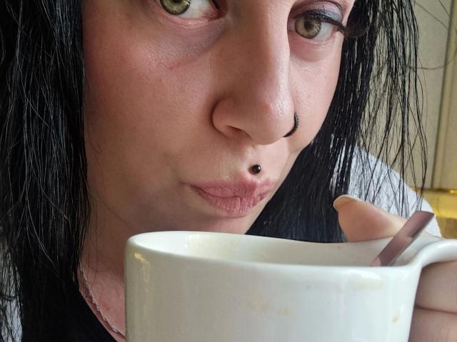 Curvy-selin-hot with black wet hair, nose ring, and lip piercing, puckers lips while holding a white mug with a brown spoon in her right hand.