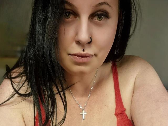 Curvy-selin-hot with black hair, nose ring, lip piercing, cross necklace, and red tank top, staring at the camera with green eyes, slightly parted lips.