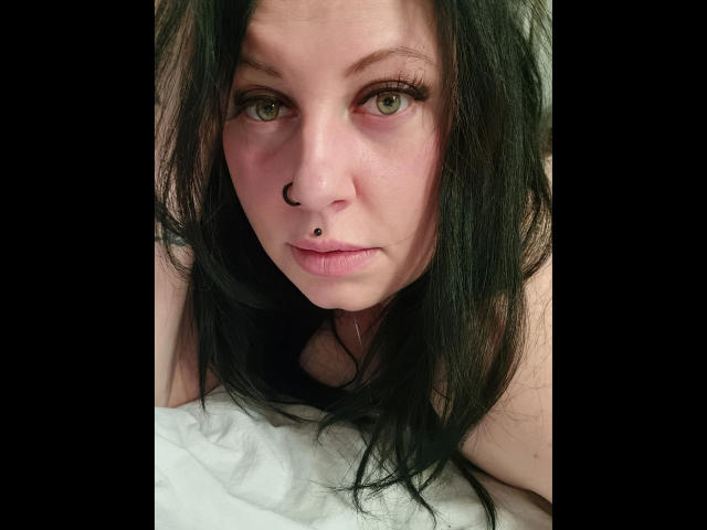 Curvy-selin-hot with black hair, green eyes, nose ring, and lip piercing, staring at the camera with a subtle smile, lying on a white surface.