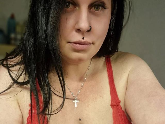 Curvy-selin-hot with black hair, nose ring, and lip piercing, wearing a red tank top and silver cross necklace, showing her bare shoulders and small nipple piercing.