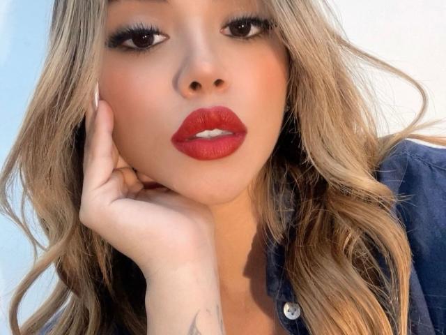 VaiolethLamour tiene lápiz labial rojo, cabello rubio ondulado y un tatuaje en la mano. Lleva una camisa azul y está tocándose la mejilla con la mano. Sus ojos están medio abiertos..