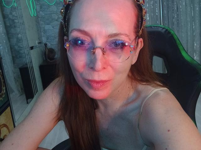 IrunchaArt with glasses, colorful headband, and pale skin, sitting in a chair, smiling, showing small tits, and a cute face, with pink and blue lighting.