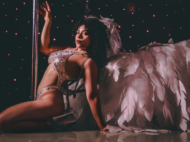 IvannaAndFriend with curly hair, wearing a glittery, revealing one-piece, sitting by a pole, arm raised, white feather wings, looking sexy, showing her tits and ass.