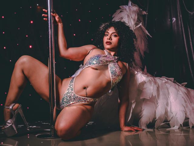 IvannaAndFriend with curly black hair, wearing a glittery, one-piece, booty-cut, sparkly bikini, and silver high heels, kneeling by a pole, with white angel wings in the background.