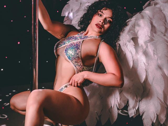IvannaAndFriend has curly black hair and wears a glittery, multicolored one-piece swimsuit that reveals her large, round breasts and curvy body. She's leaning on a silver pole with one arm, and has white, feathery angel wings on her back. Her red lipstick contrasts with her dark skin. The background is dark with small pink lights. She's looking directly at the camera with a confident expression. The composition highlights her physical features and the texture of the wings.
