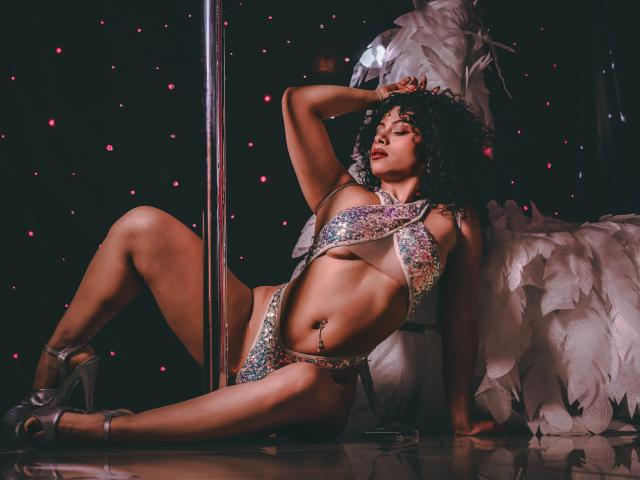 IvannaAndFriend leans against a silver pole, eyes closed, wearing a glittery, rainbow-patterned bikini that barely covers her breasts and crotch. She has curly black hair, a dark skin tone, and wears silver high heels. White feathered wings are on her right, and she has a small belly button piercing. The dark background has small red dots. She's relaxed, with one arm behind her head and the other on the pole. The lighting is low, emphasizing her body. The image has a sensual, provocative vibe.