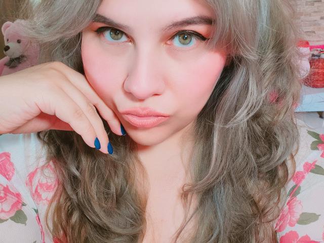 Akkanee, avec ses cheveux gris ondulés, ses ongles bleus, son blush rose et ses lèvres pulpeuses, regarde l'appareil photo avec ses yeux verts, et porte un haut fleuri. Une douce lumière rose illumine son visage..