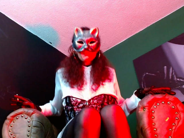 GoddessLulu wearing a cat mask, white fur-sleeved top, black vinyl corset, and red-tinted lighting. Sitting on studded leather chairs with red and green light.