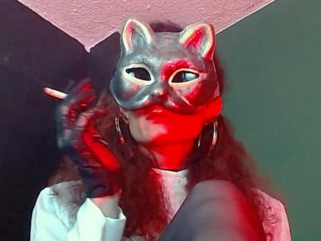 GoddessLulu wearing a cat mask, red paint on face, curly hair, gold hoop earrings, holding cigarette, white shirt, red and gray color scheme, slightly blurred photo.