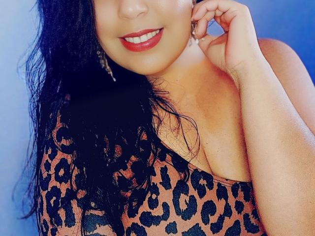 Nadjax con lunghi capelli neri, indossa una maglietta a stampa leopardo che mette in mostra il décolleté, sorride con rossetto rosso, con una mano vicino all'orecchio, indossa orecchini pendenti, sfondo blu..