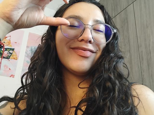 Mllevv has wavy black hair, glasses, and is sticking her tongue out with a finger pointing at her eye. She's smiling with a cute, playful expression.