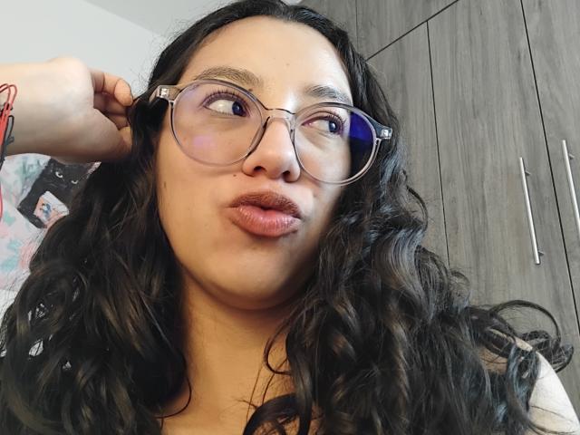 Mllevv with long black wavy hair, wearing glasses, puffy pinkish lips, brown eyes, and a black top, touching her ear, with a red bracelet on her wrist.