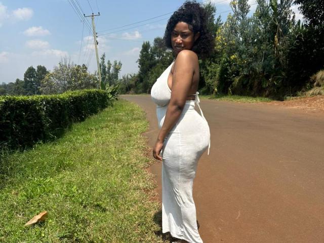 NaughtyOlivia stands on a road in a white, form-fitting dress, highlighting her curvy ass and large, round tits. She has dark curly hair and a confident smile.