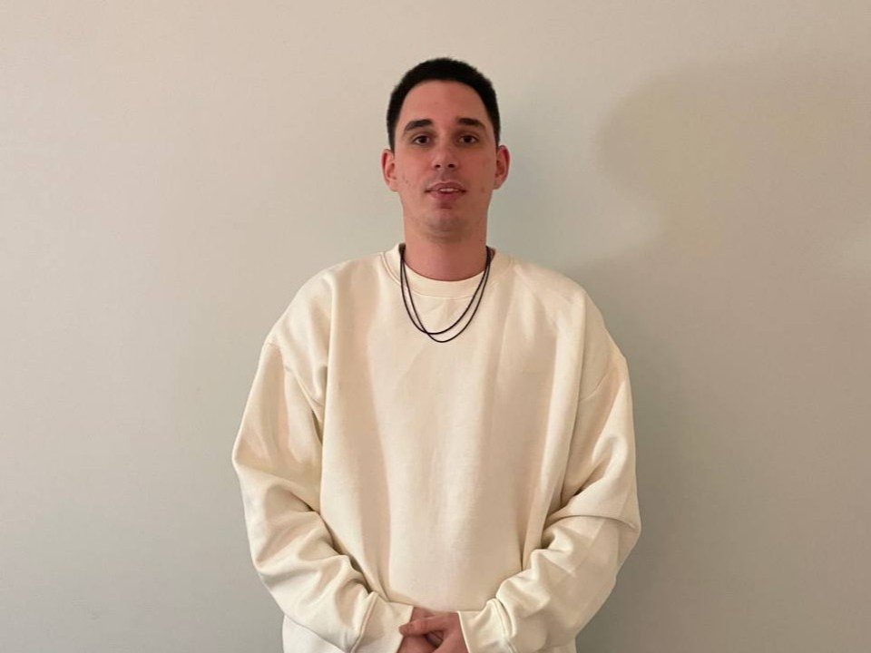 JasonSweety with short black hair, wearing a white oversized sweatshirt, black necklaces, standing against a plain wall, hands in front, neutral expression.