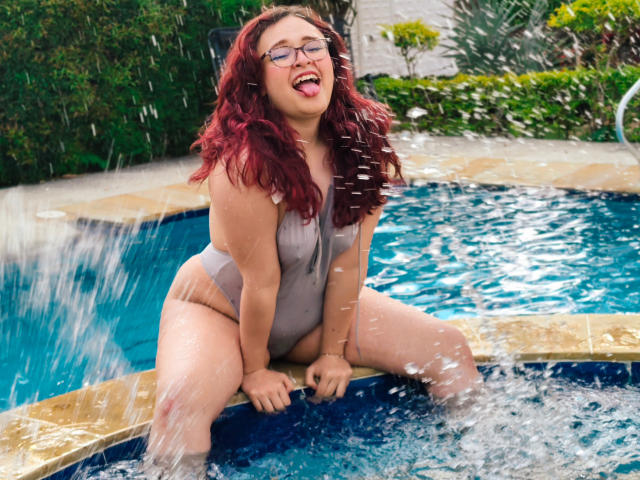 AlanisSweet with red hair, glasses, and a grey one-piece swimsuit, sitting in a blue hot tub, water splashing, tongue out, hands on edge, curvy, showing her ass.