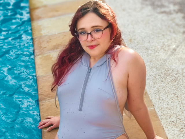AlanisSweet, with red hair and glasses, wears a wet, gray, zip-up one-piece, showing her tits, by the blue pool, sitting on stone, smiling slightly.